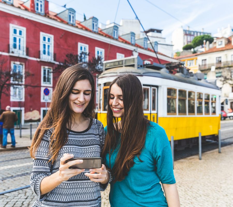 Two girls in Lisbon with the famous yellow tram on background. Young happy women looking at a smartphone in the city. Lifestyle and travel concepts.